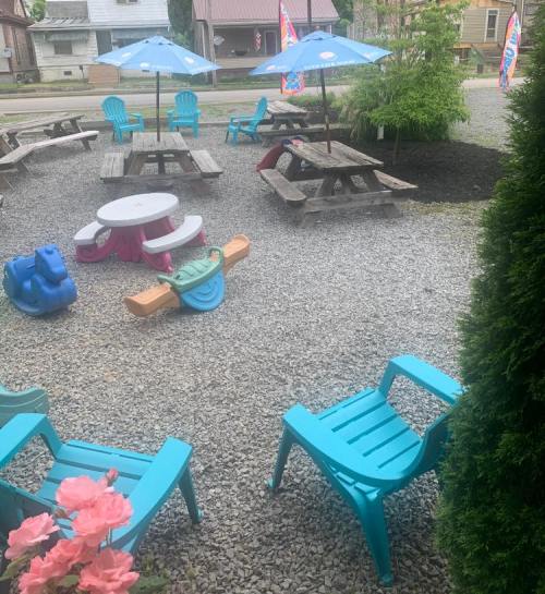 Outdoor seating area with blue chairs, picnic tables, umbrellas, and children's play equipment on gravel.