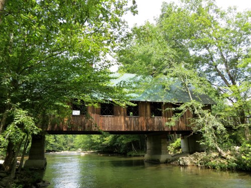 A wooden covered bridge spans a serene river, surrounded by lush green trees and a clear blue sky.