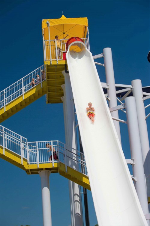 A child slides down a large water slide, with a lifeguard tower and spectators in the background under a clear blue sky.