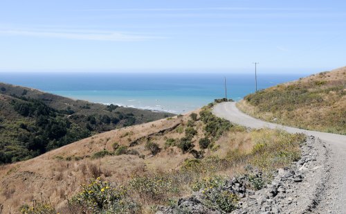 A winding dirt road leads down a hillside to the ocean, with blue waters and a clear sky in the background.