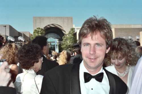 A man in a tuxedo smiles at the camera, surrounded by a crowd at an outdoor event with a modern building in the background.