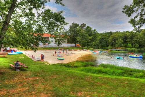 A sandy beach by a lake with people enjoying activities, surrounded by trees and colorful boats.