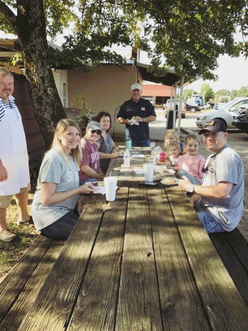 A group of people enjoying a meal at an outdoor table under a tree, smiling and chatting together.
