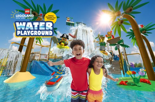 Two children joyfully pose in front of a colorful LEGO water playground with slides and waterfalls under a sunny sky.