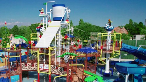 A colorful water park with slides, pools, and play structures surrounded by trees and blue skies.