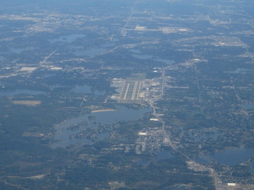 Aerial view of a landscape featuring lakes, a small airport, and urban areas in the distance.