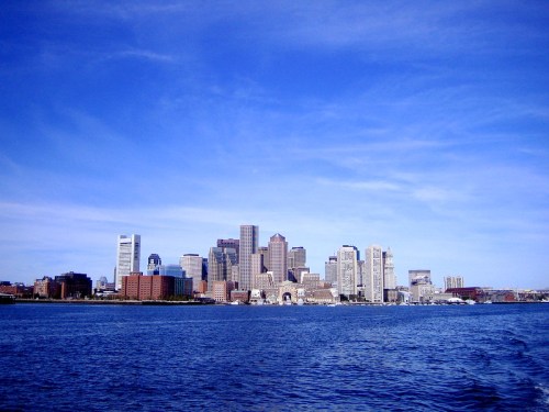 A skyline view of a city with tall buildings against a clear blue sky, reflecting on the water in the foreground.