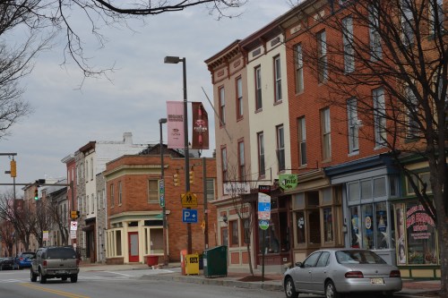 A street view of a small town with brick buildings, shops, and parked cars under a cloudy sky.