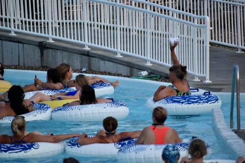 A group of people relax in inner tubes in a pool, with one person raising their hand, possibly holding something.