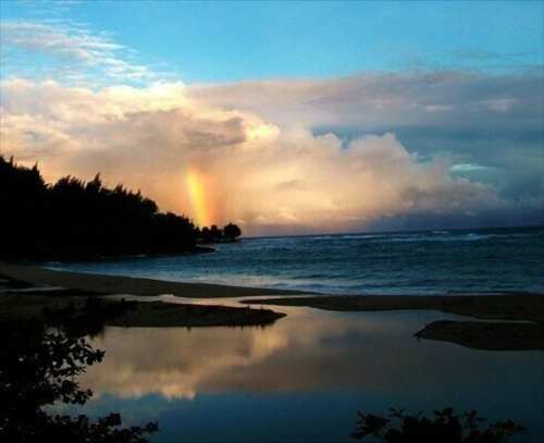 A serene beach scene with a rainbow in the sky, reflecting in a calm pool of water at sunset.
