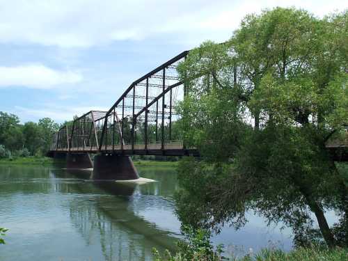 A historic metal bridge spans a calm river, surrounded by lush greenery and a clear blue sky.