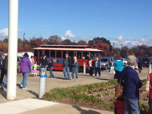 A crowd of people gathers near a red trolley on a sunny day, with colorful autumn trees in the background.