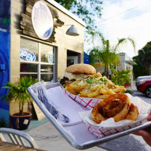 A tray holding a burger and fries, with an onion ring, in front of a colorful restaurant with an "Open" sign.