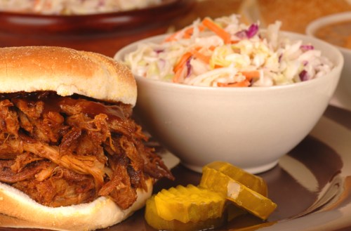 A pulled pork sandwich with barbecue sauce, served with coleslaw and pickles on a plate.