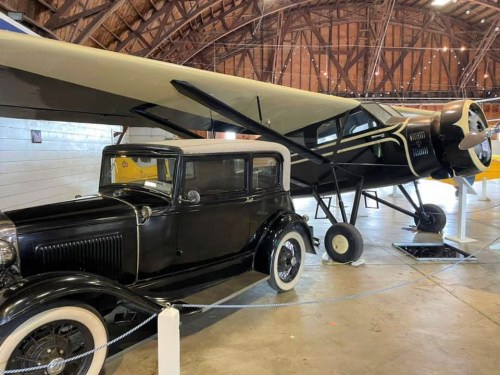 A vintage black car parked next to a classic airplane inside a large wooden hangar.