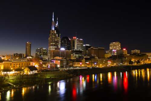 Nashville skyline at night, featuring illuminated buildings and reflections on the water.