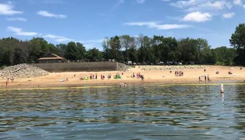 A sandy beach with people enjoying the sun, surrounded by trees and a rocky area in the background under a blue sky.