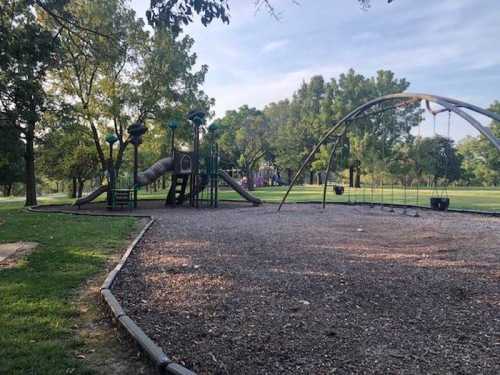 A playground with a slide and swings, surrounded by trees and grassy areas on a sunny day.