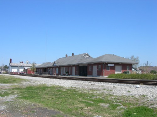 A brick train station with a gray roof, set against a clear blue sky and surrounded by grass and railroad tracks.