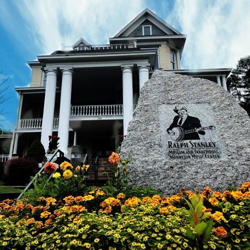 A stone monument with a musician engraving in front of a large historic building, surrounded by colorful flowers.