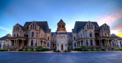 Historic stone mansion with intricate architecture, featuring a central tower and landscaped grounds under a colorful sky.