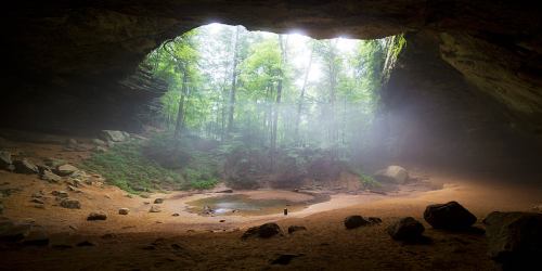 A serene cave opening reveals a misty forest with sunlight filtering through, illuminating a sandy floor and a small pond.