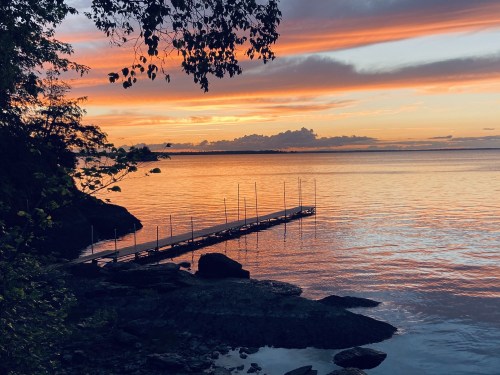 A serene sunset over calm waters, with a wooden dock extending into the lake and trees framing the scene.