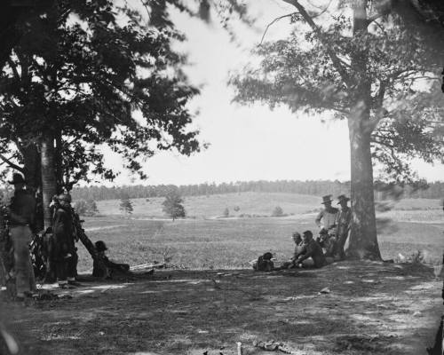 A historical black-and-white photo of soldiers resting under trees in a field, with a distant landscape visible.