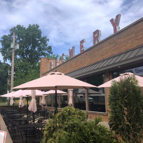 Exterior of a brewery with pink umbrellas on patio tables and a sign reading "Brewery" against a cloudy sky.
