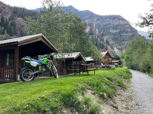A green dirt bike parked near wooden cabins, surrounded by trees and mountains in a scenic outdoor setting.