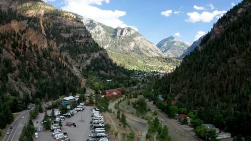 Aerial view of a valley surrounded by mountains, featuring a river, trees, and a parking area for RVs.