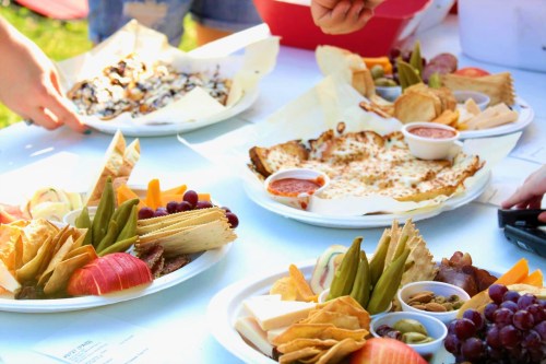 A variety of colorful platters with fruits, cheeses, crackers, and dips on a table outdoors.