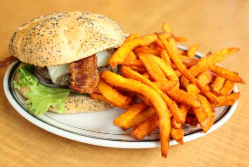 A poppy seed bun burger with bacon, cheese, and lettuce, served with a side of crispy sweet potato fries.