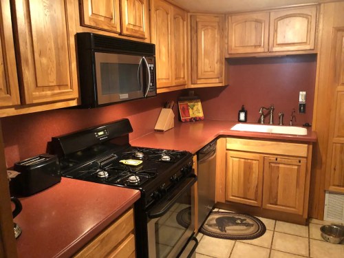 A cozy kitchen with wooden cabinets, a black stove, microwave, and a sink on a red countertop. Tile flooring is visible.
