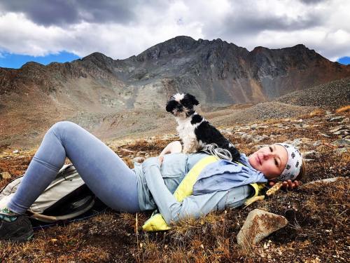 A woman relaxes on rocky terrain with a small dog beside her, mountains in the background under a cloudy sky.