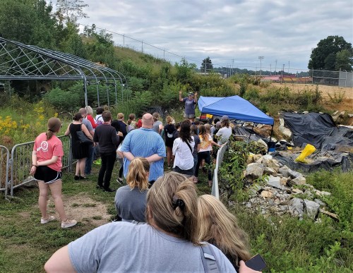 A group of people gathers outdoors, listening to a speaker near a blue tent and overgrown vegetation.