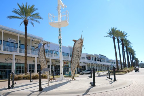 A sunny outdoor shopping area featuring large fish sculptures and palm trees, with shops lining the background.