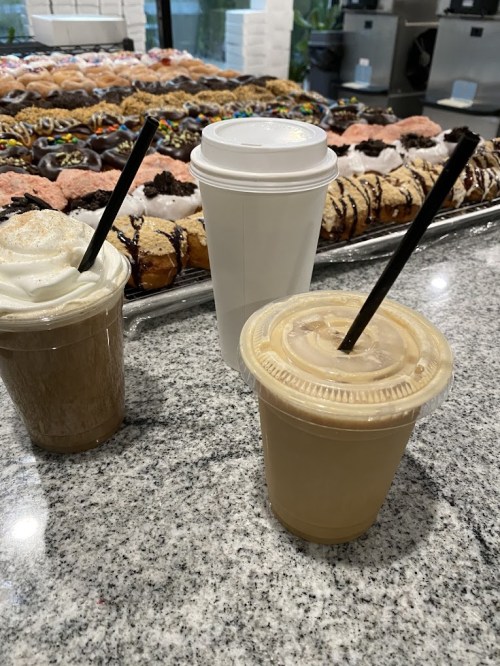 Three coffee drinks in front of a display of assorted donuts on a marble countertop.