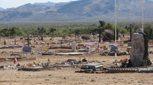 A desert cemetery with gravestones, flowers, and flags, surrounded by mountains and sparse vegetation.
