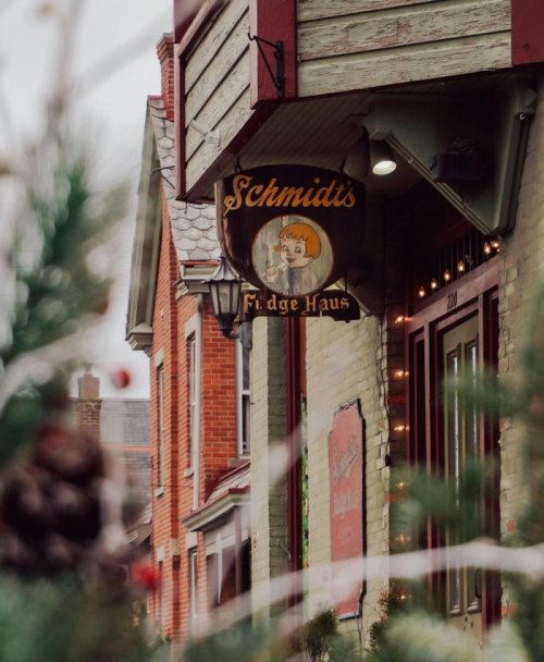 Sign for "Schmidt's Fudge Haus" hanging outside a charming brick building, surrounded by festive greenery.