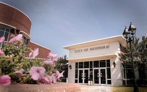 Entrance of the City of Bedford building, featuring modern architecture and blooming flowers in the foreground.