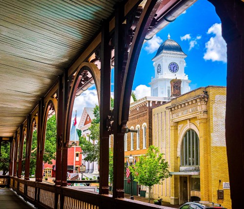 View of a town square with a clock tower, framed by an arched wooden walkway and blue sky with clouds.