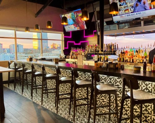 A modern bar with a stylish design, featuring a long counter, stools, and large windows showcasing a city view.