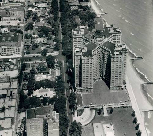 Aerial view of a large building near the shoreline, surrounded by trees and residential areas. Waves visible in the water.