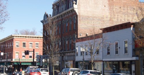 A street view featuring historic buildings, shops, and parked cars under a clear blue sky.