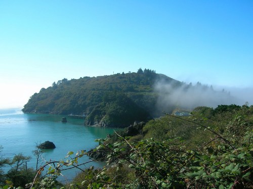 A serene coastal landscape featuring a misty hill, clear blue water, and lush greenery under a bright sky.