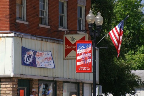 A street view featuring a Budweiser sign, a Miller Lite banner, and a "Welcome to Kirkland" flag with fireworks design.