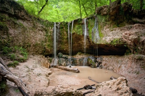 A serene waterfall cascades over rocky terrain into a calm pool, surrounded by lush green trees and earthy tones.