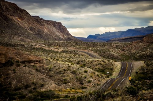 Winding road through a rugged desert landscape with mountains under a cloudy sky.