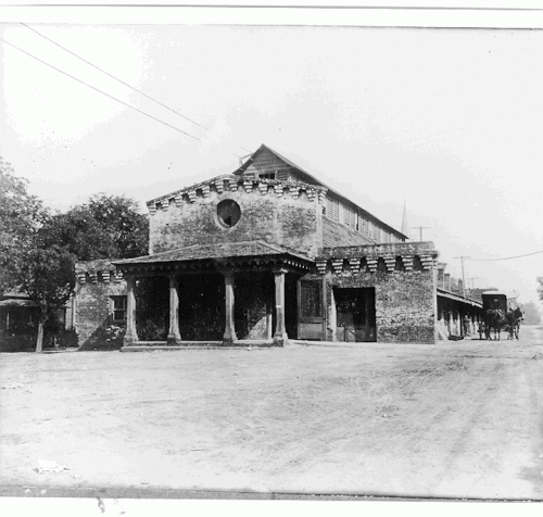 Historic black-and-white photo of a brick building with a porch, surrounded by trees and a dirt road.